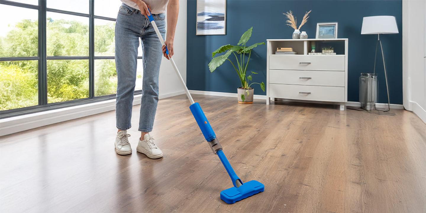 Person, die einen Holzboden in einem modernen Raum mit blauen Wänden, weißem Schrank, Pflanzen und großen Fenstern mit Blick auf eine grüne Landschaft wischt.