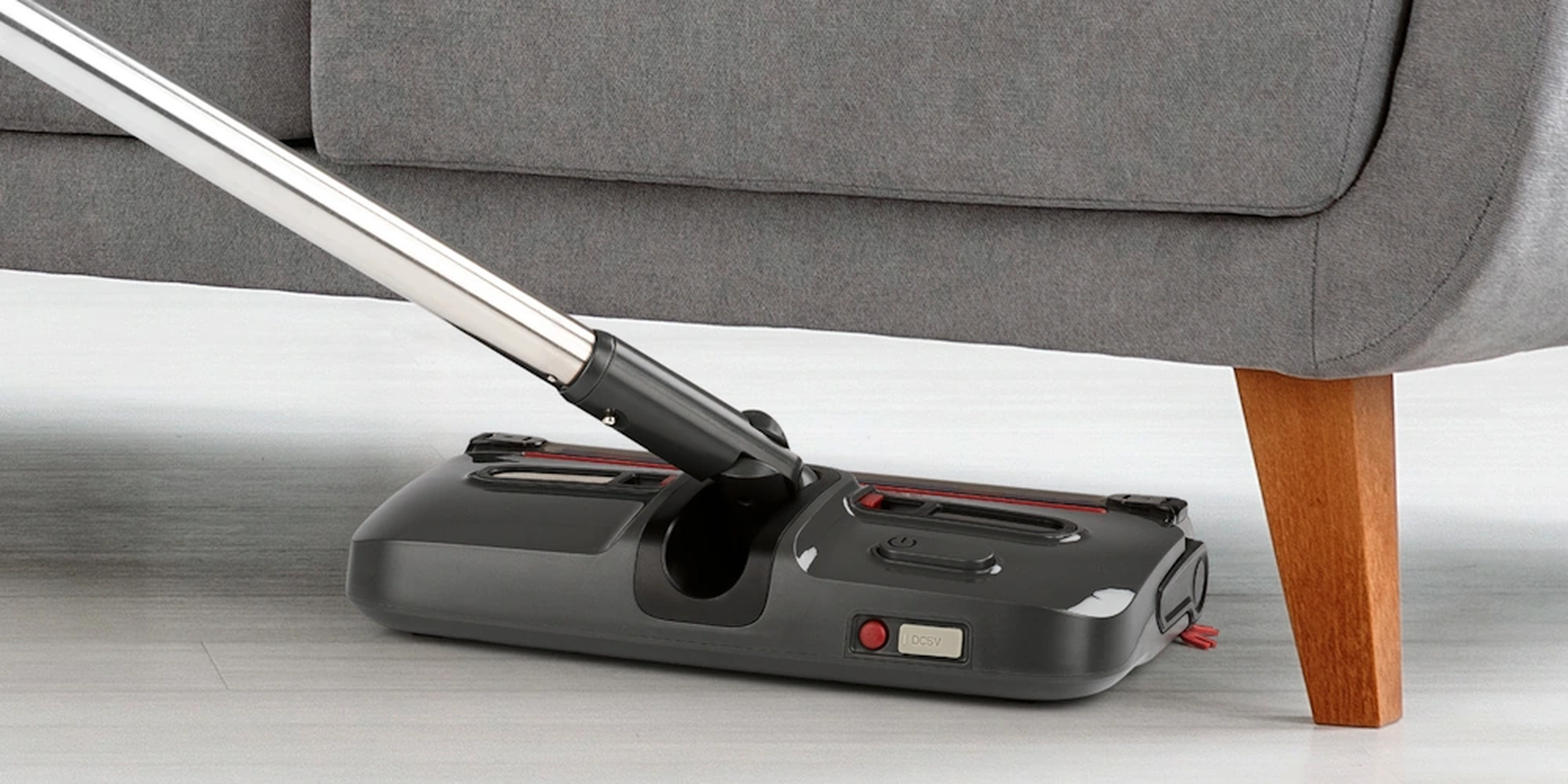 Ein kabelloser Staubsauger, der unter ein graues Sofa greift und dabei sein schlankes, flaches Design zeigt, das eine einfache Reinigung in engen Räumen ermöglicht.