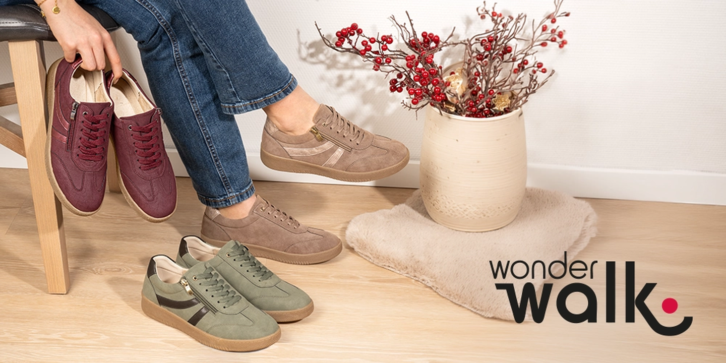 Eine Person sitzt auf einem Stuhl, trägt beige Turnschuhe, hält ein Paar burgunderrote Schuhe in der Hand, auf dem Boden stehen grüne Turnschuhe, daneben steht eine Vase mit roten Beeren.