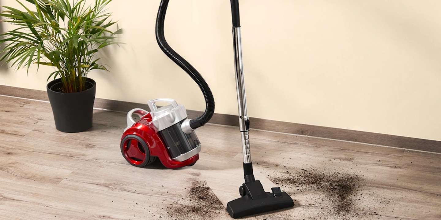 Ein roter Staubsauger mit einem flexiblen Schlauch auf einem Holzboden, neben verstreutem Schmutz. In der Nähe steht eine Topfpflanze.