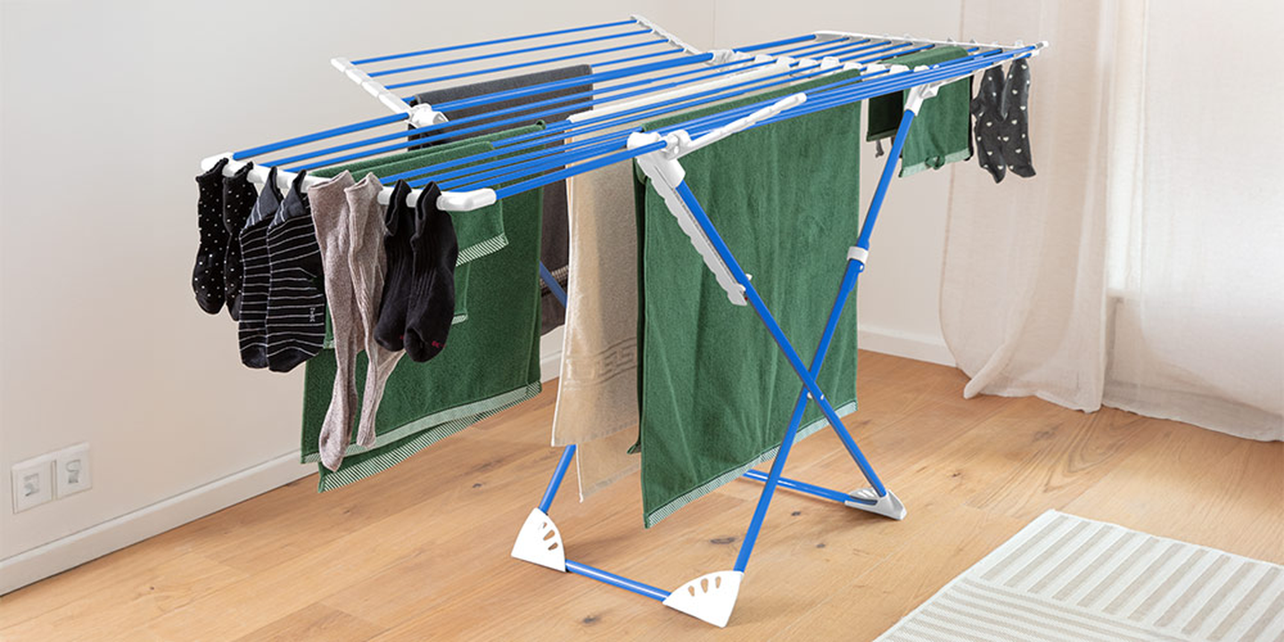 Auf einem blauen Klappgestell trocknen Kleider, Handtücher und Socken in einem hellen Raum mit Holzboden und Teppich.