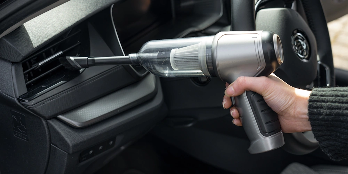 Eine Hand, die einen kleinen Staubsauger hält und die Lüftungsdüse im Innenraum eines Autos reinigt.