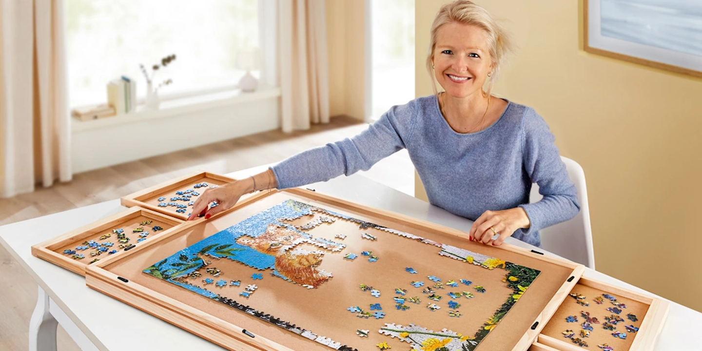 Eine Frau in einem blauen Pullover lächelt, während sie in einem hellen Raum auf einem hölzernen Puzzletisch mit Sortierfächern ein Puzzle zusammensetzt.