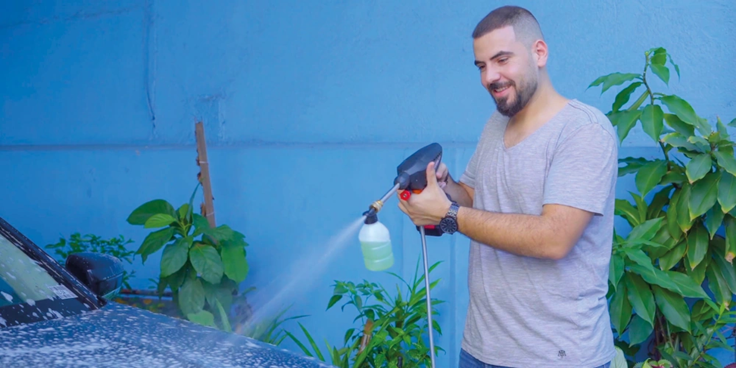 Ein Mann wäscht vor einer blauen Wand, umgeben von viel Grün, ein Auto mit einem Hochdruckreiniger.