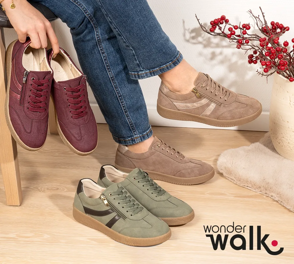 Eine Person sitzt auf einem Stuhl, trägt beige Turnschuhe, hält ein Paar burgunderrote Schuhe in der Hand, auf dem Boden stehen grüne Turnschuhe, daneben steht eine Vase mit roten Beeren.