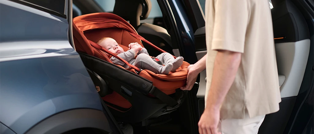 Eine Person setzt ein Baby in einem roten Kindersitz in ein Auto. Das Baby trägt graue Kleidung und wirkt entspannt und geborgen.