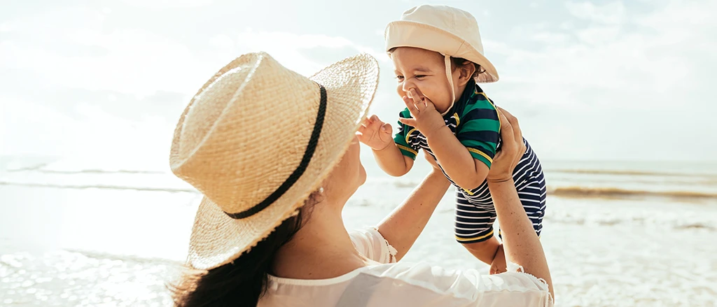 Eine Frau mit Sonnenhut hebt ein lächelndes Baby, das ebenfalls einen Hut und ein gestreiftes Outfit trägt, vor einer sonnigen Strandkulisse hoch.