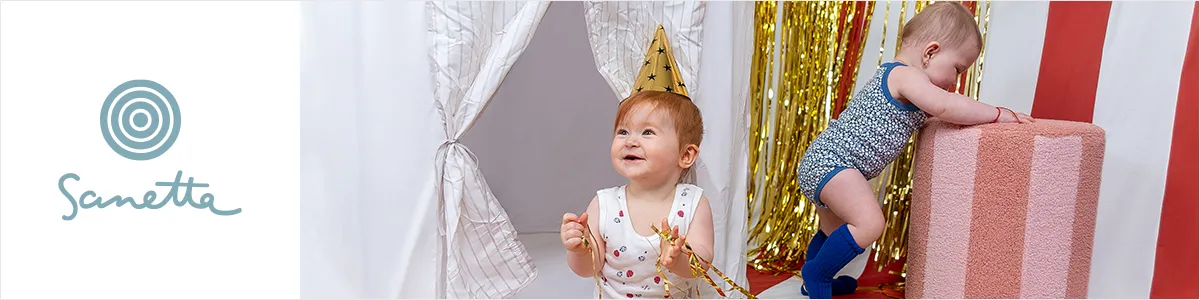 Zwei Kleinkinder spielen; eines sitzt mit einem goldenen Kegelhut auf dem Kopf, das andere steht neben einem gestreiften Kissen mit glänzenden Luftschlangen. An der Seite ist das „Sanetta“-Logo zu sehen.