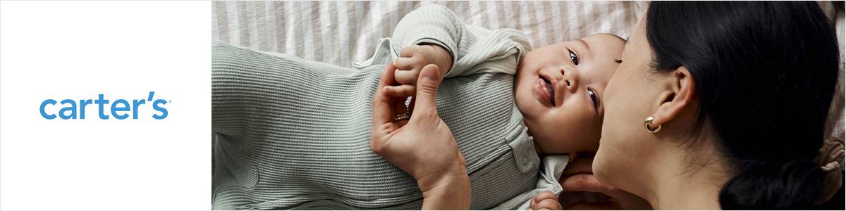 Ein lächelndes Baby in einem hellgrünen Outfit liegt auf einem Bett und hält die Hand einer Frau neben dem Carter's-Logo.