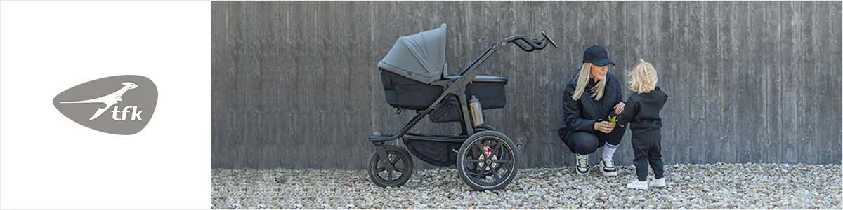 Eine kniende Frau interagiert mit einem Kind neben einem schwarzen Kinderwagen vor einer grauen Wand; beide sind schwarz gekleidet. Links ist das TFK-Logo zu sehen.