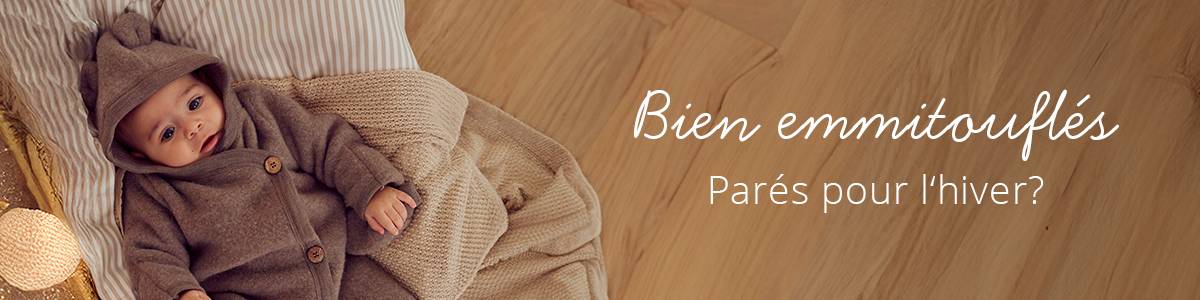 Bébé vêtu d'une confortable tenue marron avec des oreilles d'ours, allongé sur une couverture moelleuse. Le texte indique « Bien emmitouflés. Parés pour l'hiver ? »