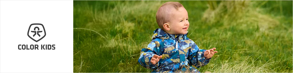 Ein Baby in einer bunten Jacke sitzt auf dem Rasen, lächelt und schaut zur Seite. Links ist das „Color Kids“-Logo zu sehen.