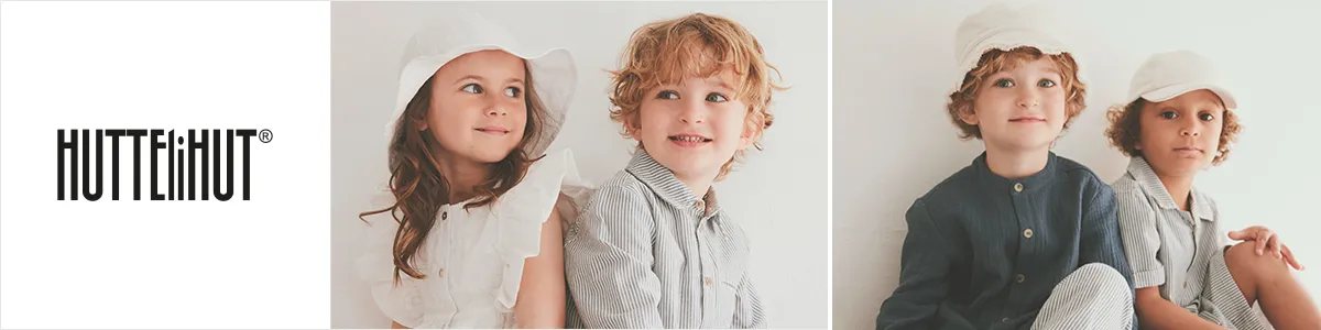 Vier kleine Kinder in weißen und grauen Outfits und Mützen, die lächelnd vor einem weißen Hintergrund sitzen, auf dem links der Schriftzug „Huttelihut“ zu sehen ist.