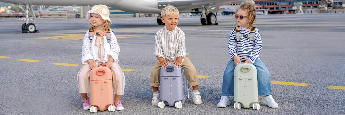 Drei Kinder sitzen auf pastellfarbenen Koffern an einem Flughafen, mit einem Flugzeug im Hintergrund.