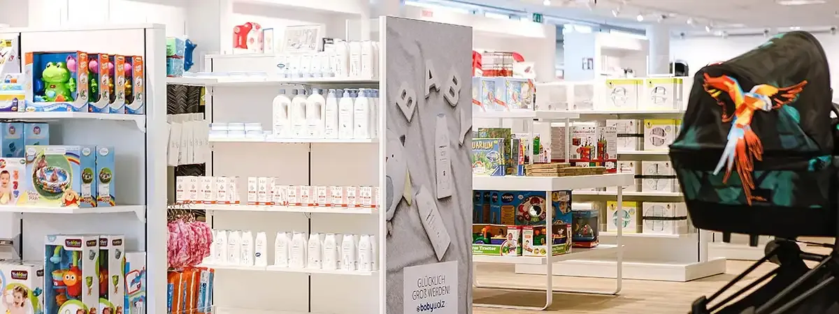 Innenansicht einer baby-walz Filiale mit Regalen voller Babyprodukte, Pflegeartikel, Spielzeug und Zubehör. Im Vordergrund ein Kinderwagen mit buntem Papageienmotiv auf dem Verdeck. Im Hintergrund gut beleuchtete Regale mit verschiedenen Produkten für Babys und Kleinkinder
