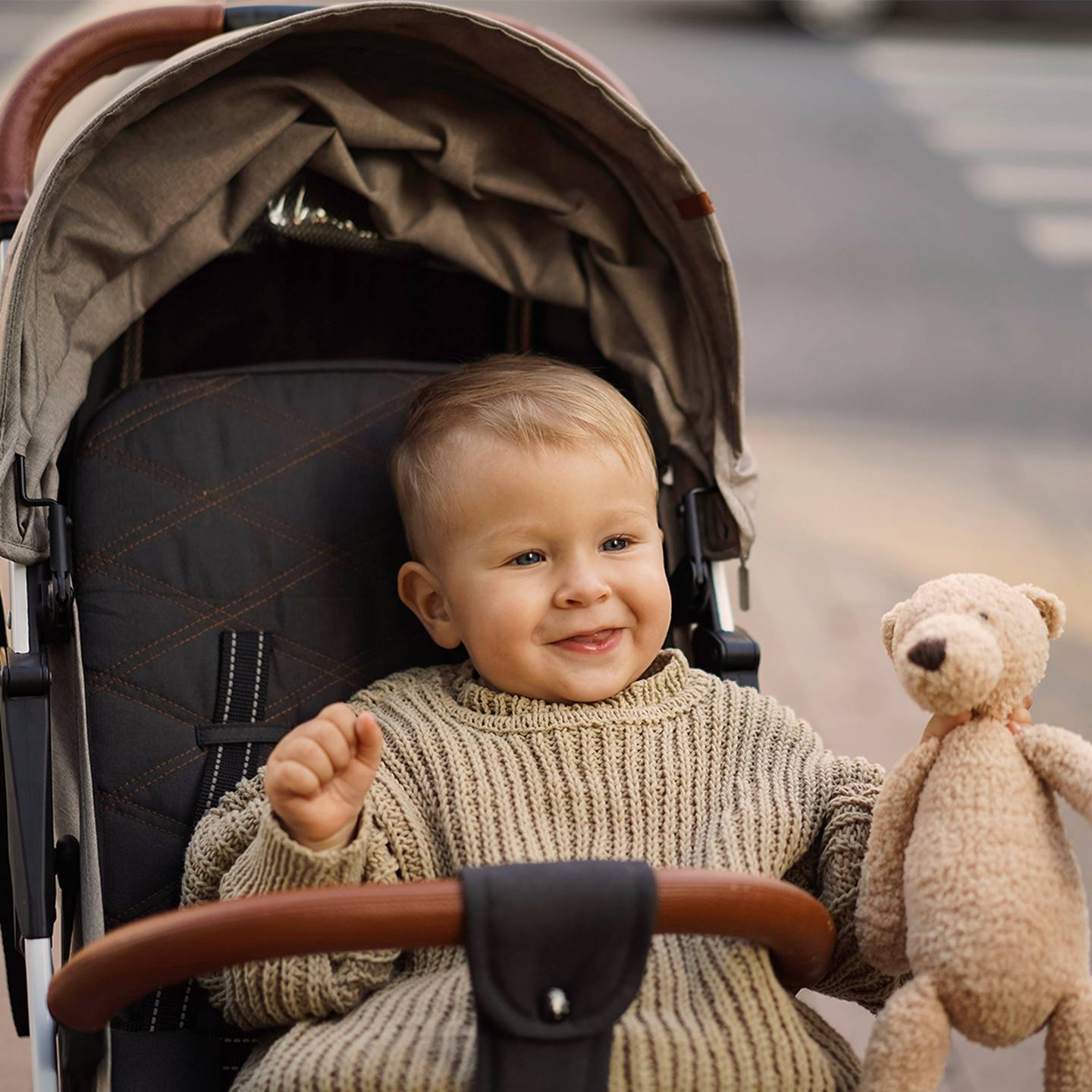 Ein lächelndes Kleinkind in einem Kinderwagen hält einen Stoffbären. Das Kind trägt einen gestrickten Pullover. Der Hintergrund zeigt einen Zebrastreifen und eine Straße.