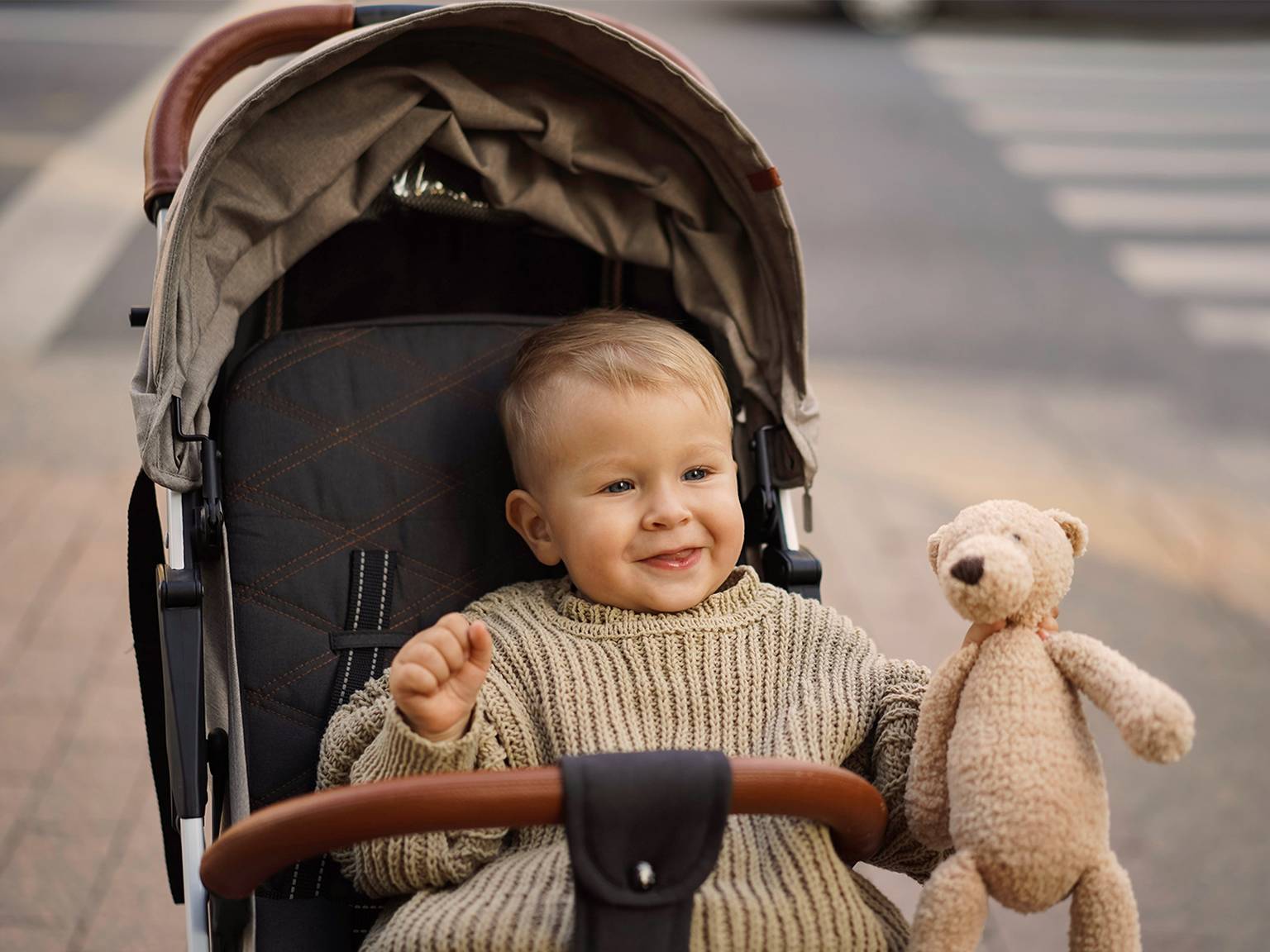 Ein lächelndes Kleinkind in einem Kinderwagen hält einen Stoffbären. Das Kind trägt einen gestrickten Pullover. Der Hintergrund zeigt einen Zebrastreifen und eine Straße.