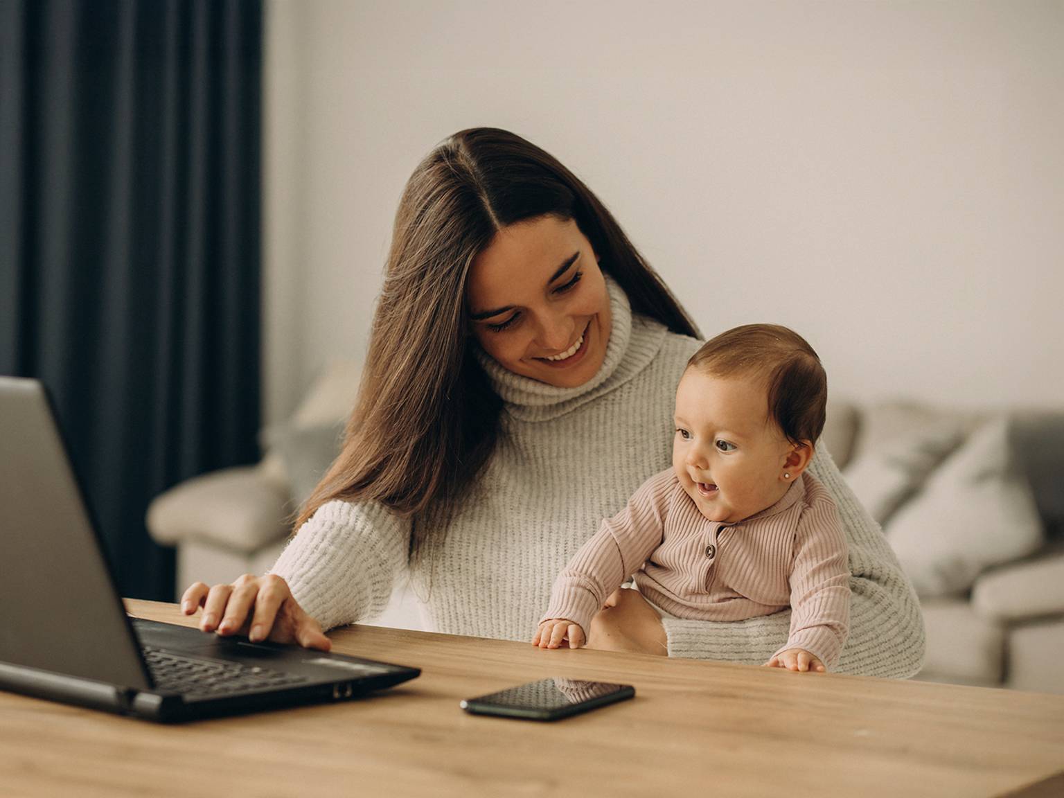 Eine Frau lächelt, während sie ein Baby hält und an einem Tisch einen Laptop benutzt. In der Nähe liegt ein Smartphone. Die Umgebung ist ein gemütlicher, moderner Raum.