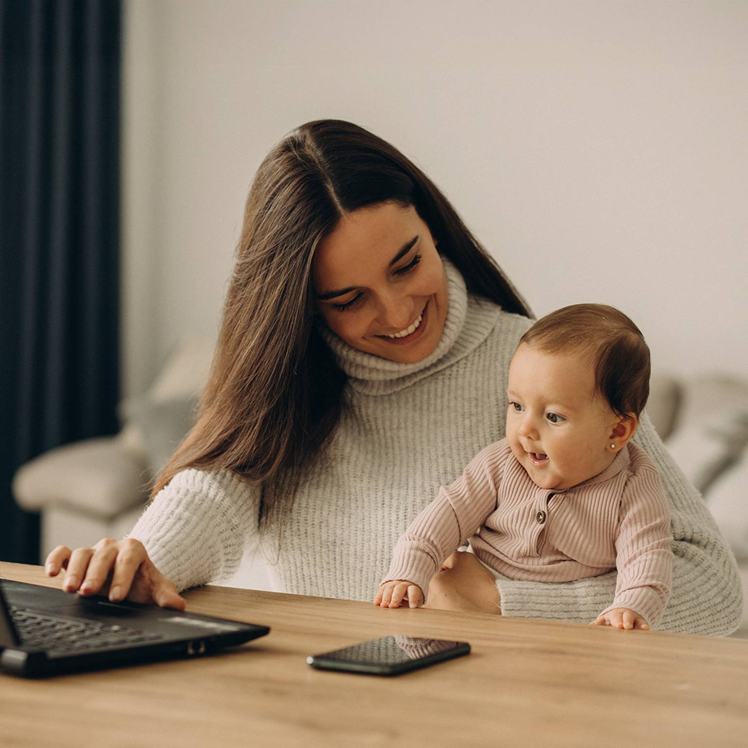 Eine Frau lächelt, während sie ein Baby hält und an einem Tisch einen Laptop benutzt. In der Nähe liegt ein Smartphone. Die Umgebung ist ein gemütlicher, moderner Raum.