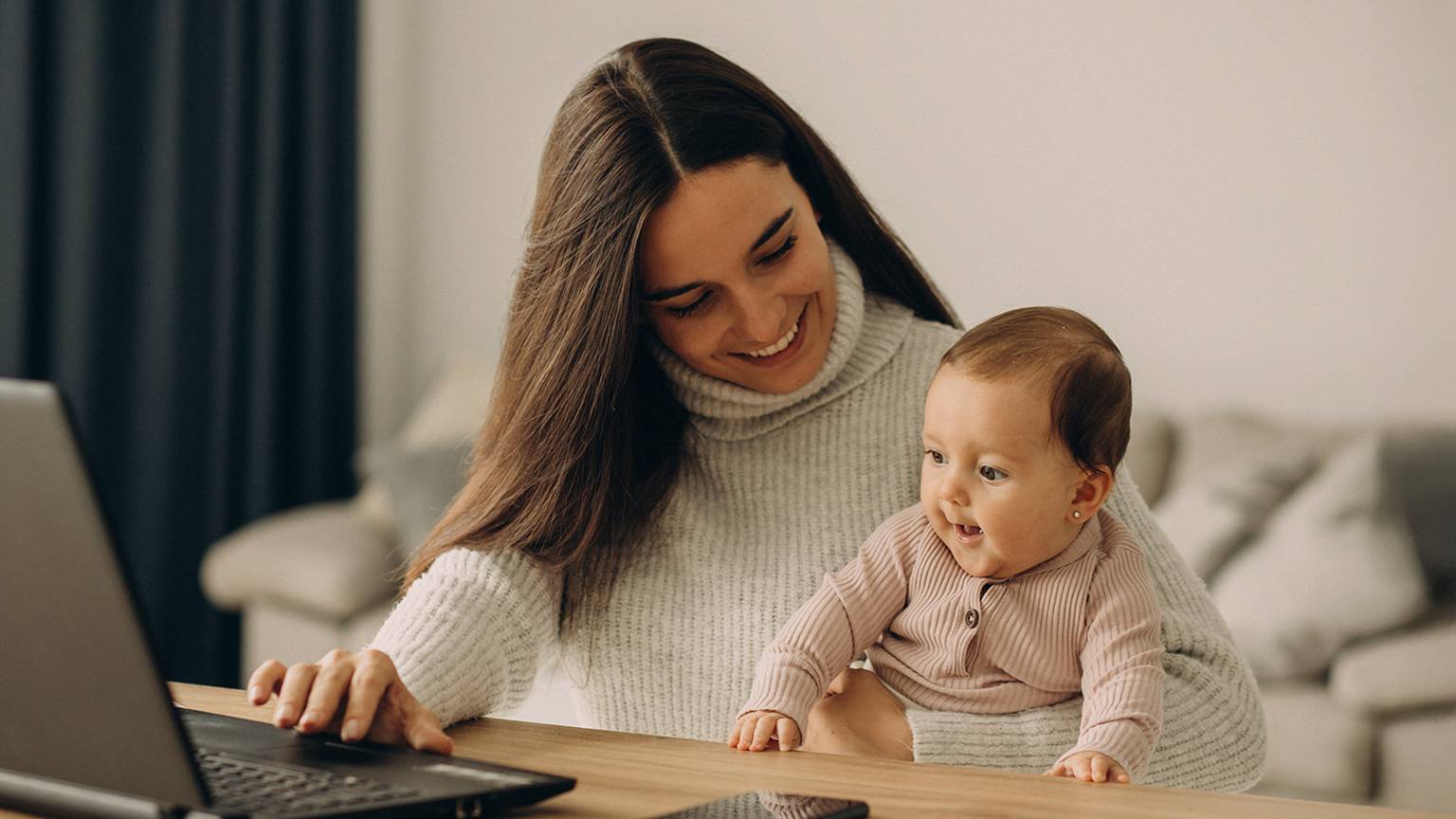 Eine Frau lächelt, während sie ein Baby hält und an einem Tisch einen Laptop benutzt. In der Nähe liegt ein Smartphone. Die Umgebung ist ein gemütlicher, moderner Raum.