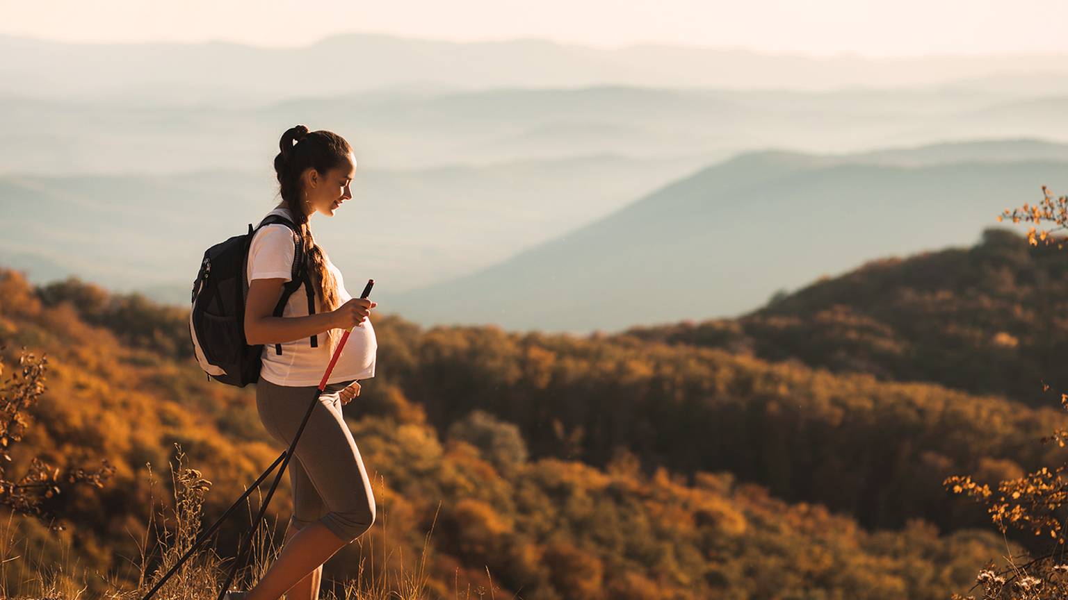 Schwangere Frau wandert mit Trekkingstöcken und genießt die malerische Aussicht auf Hügel und Herbstlaub bei einem warmen Sonnenuntergang.