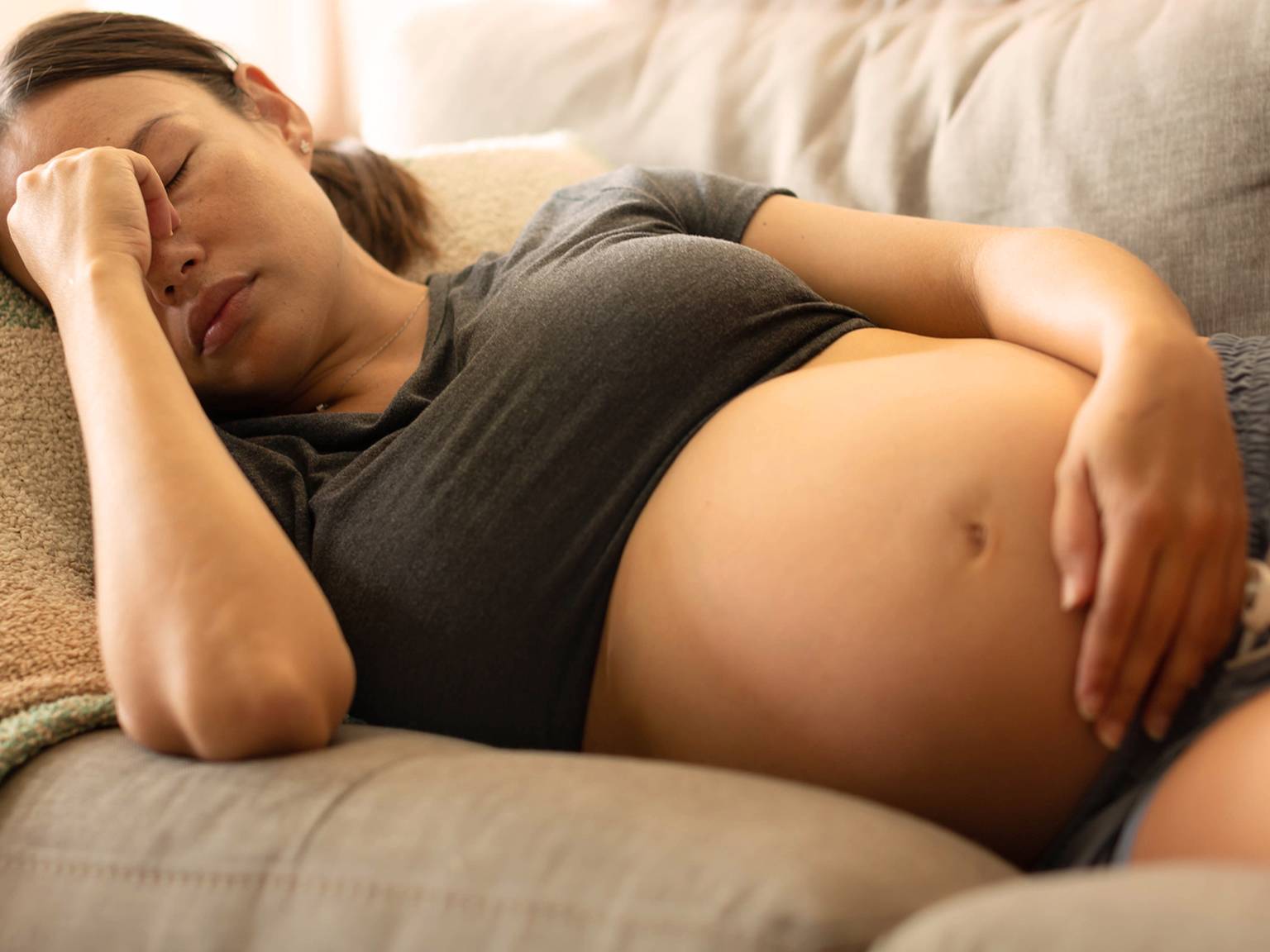 Schwangere Frau ruht auf einer Couch. Sie trägt ein dunkles T-Shirt und Shorts und hat eine Hand auf der Stirn und die andere auf dem Bauch.