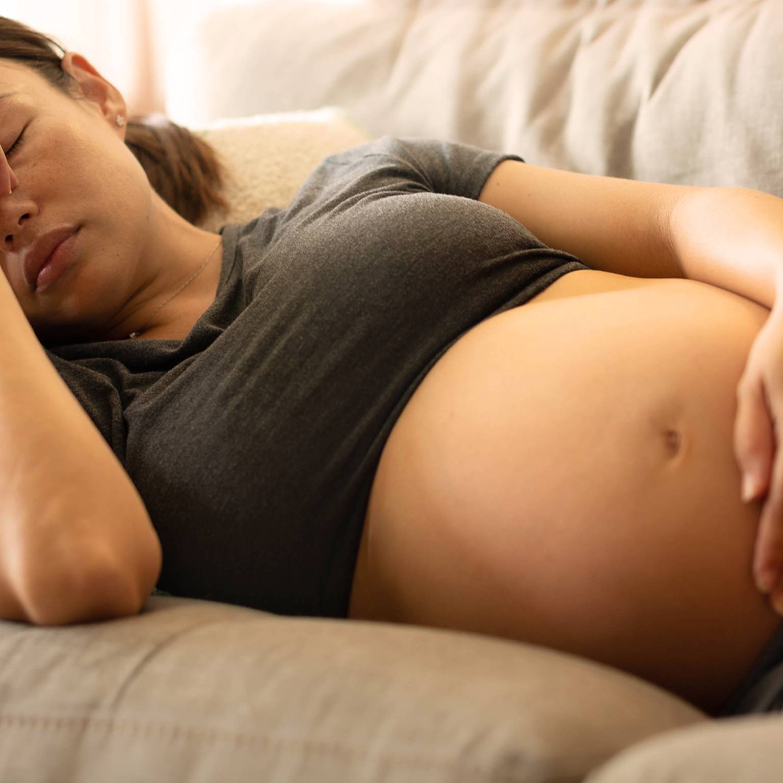 Schwangere Frau ruht auf einer Couch. Sie trägt ein dunkles T-Shirt und Shorts und hat eine Hand auf der Stirn und die andere auf dem Bauch.