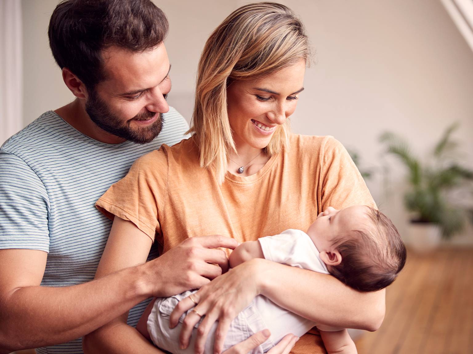 Frau hält lächelnd kleines baby auf dm Arm und der Mann steht hinter ihr und lächelt