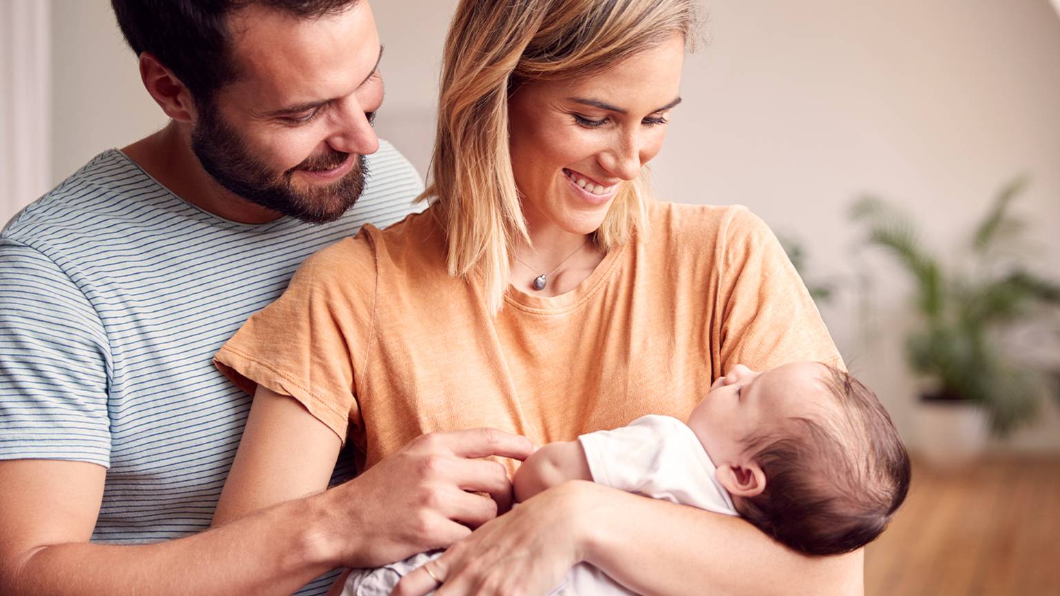 Frau hält lächelnd kleines baby auf dm Arm und der Mann steht hinter ihr und lächelt