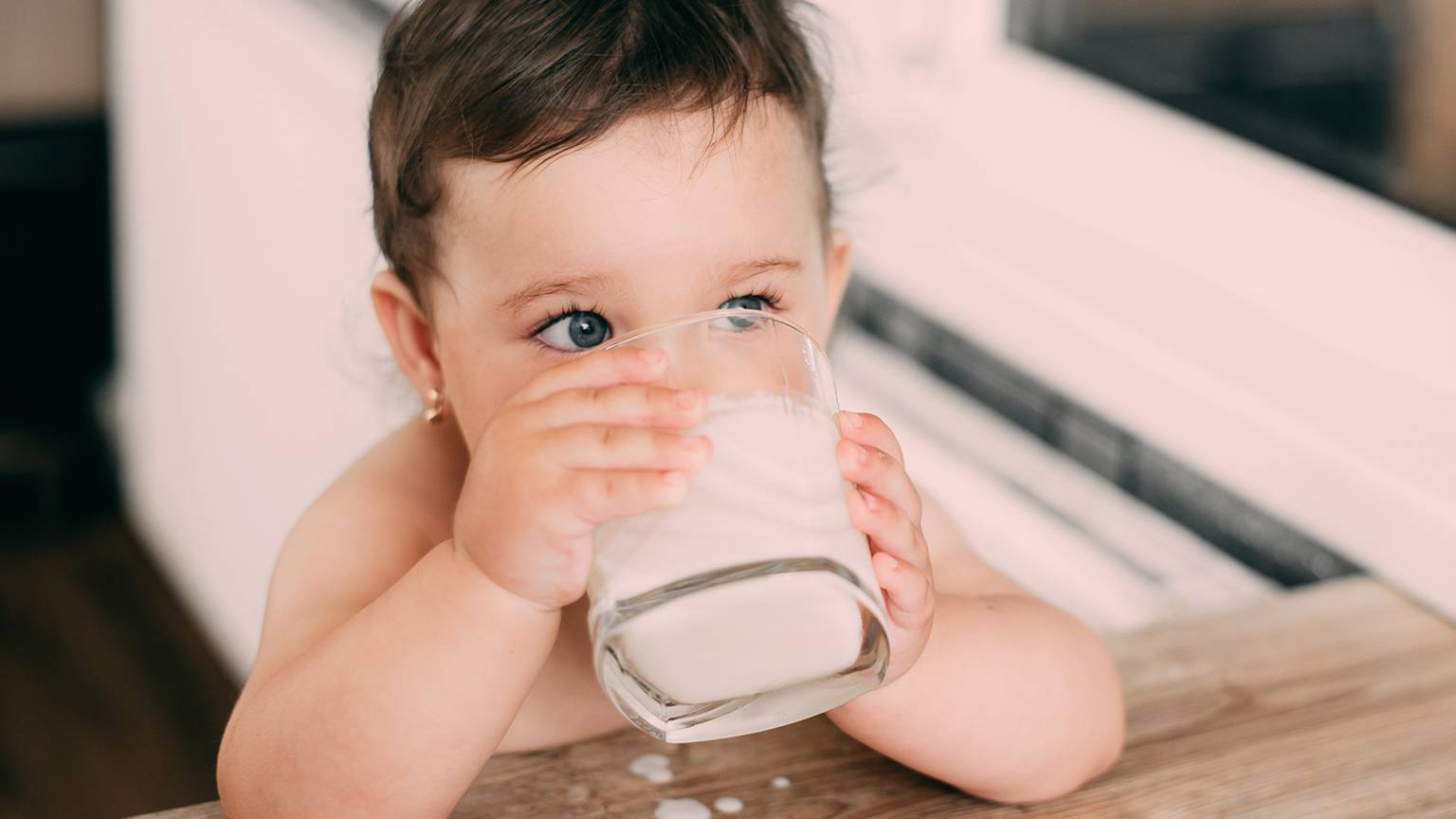 Ein Kleinkind mit kurzen dunklen Haaren trinkt Milch aus einem Glas an einem Holztisch, wobei etwas Milch auf die Tischoberfläche verschüttet ist.