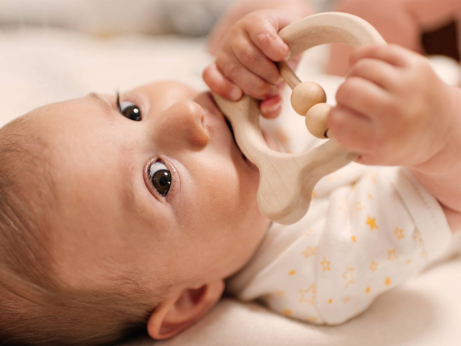 Das Baby liegt, trägt einen Strampler mit Sternenmuster, hält ein hölzernes Beißspielzeug und kaut darauf herum, während es mit großen Augen nach oben blickt.