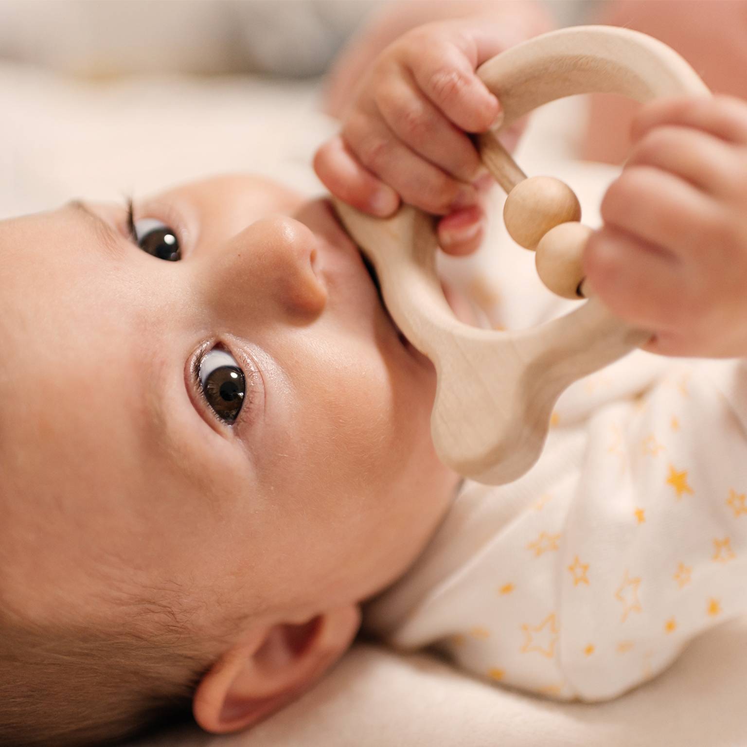 Das Baby liegt, trägt einen Strampler mit Sternenmuster, hält ein hölzernes Beißspielzeug und kaut darauf herum, während es mit großen Augen nach oben blickt.