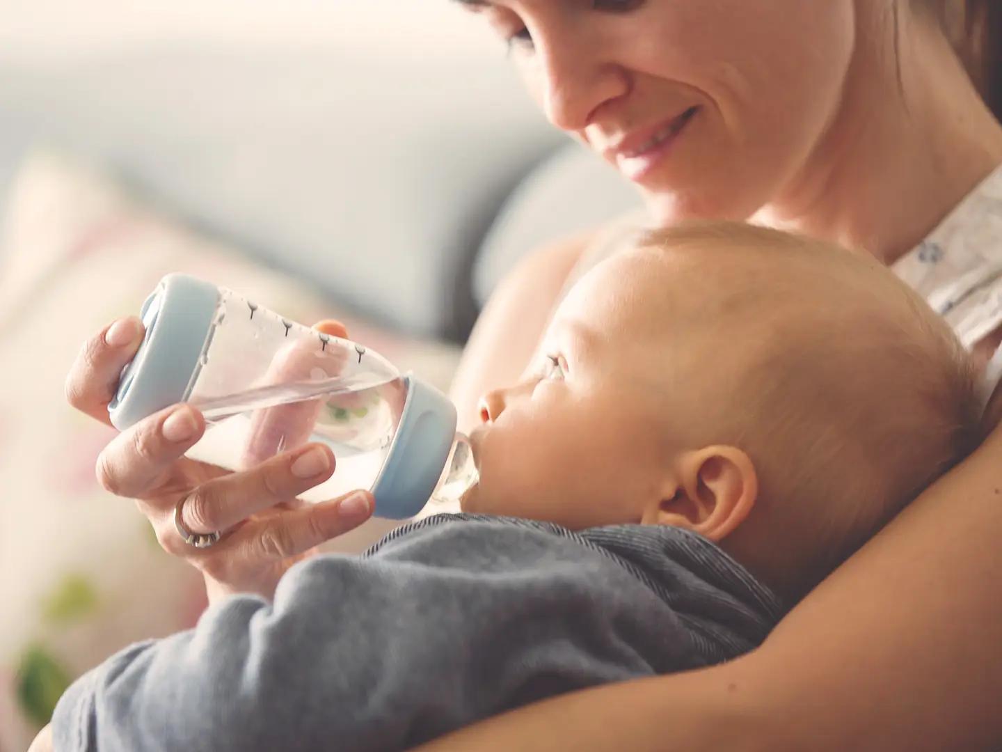 Eine lächelnde Frau hält ein Baby im Arm und füttert es mit der Flasche. Das Baby trägt ein blaues Outfit.