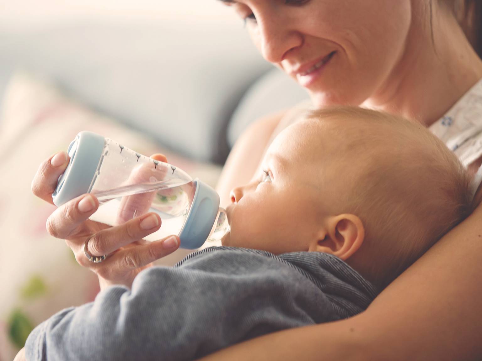 Eine lächelnde Frau hält ein Baby im Arm und füttert es mit der Flasche. Das Baby trägt ein blaues Outfit.