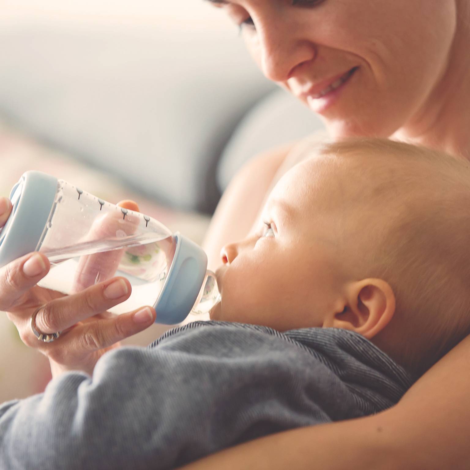 Eine lächelnde Frau hält ein Baby im Arm und füttert es mit der Flasche. Das Baby trägt ein blaues Outfit.