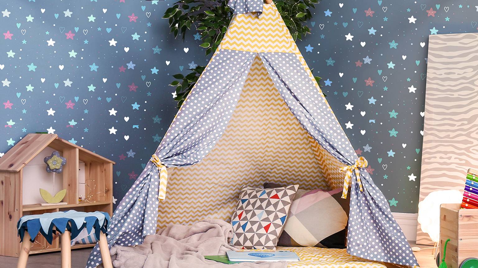 Kinderspielzimmer mit einem gelb-grau gepunkteten Tipi, bunten Kissen, einem kleinen Holztisch und einer Tapete mit Sternen.