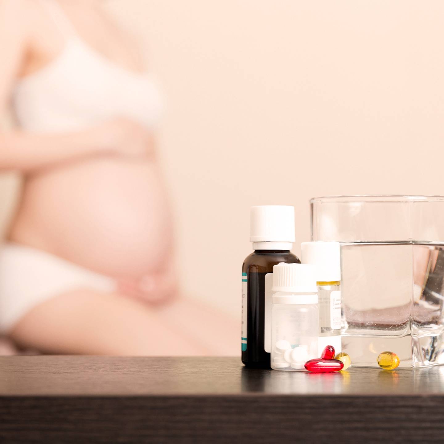 Im Hintergrund eine schwangere Frau; im Vordergrund sind ein Glas Wasser und verschiedene Medizinfläschchen und Pillen auf einem Tisch zu sehen.