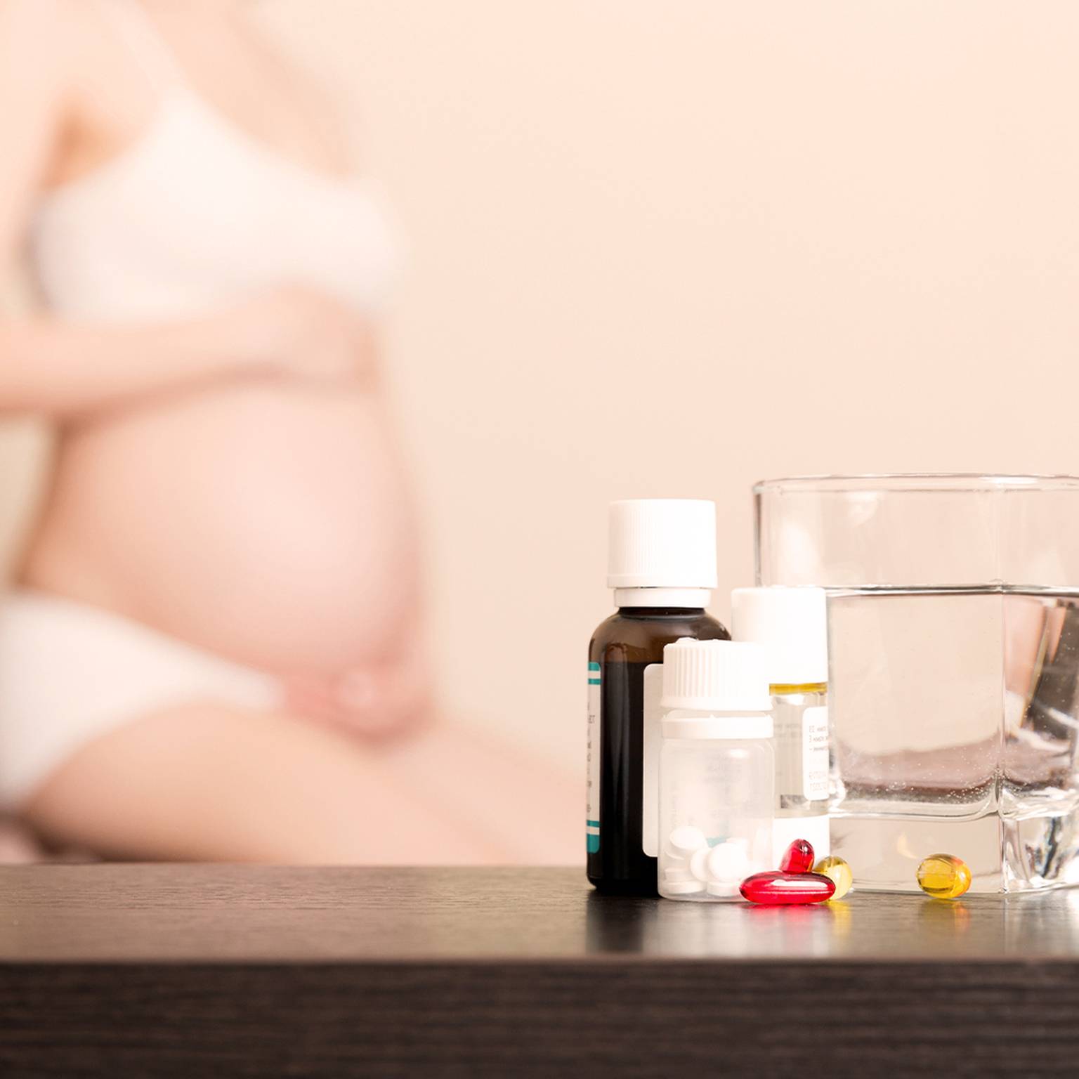 Im Hintergrund eine schwangere Frau; im Vordergrund sind ein Glas Wasser und verschiedene Medizinfläschchen und Pillen auf einem Tisch zu sehen.