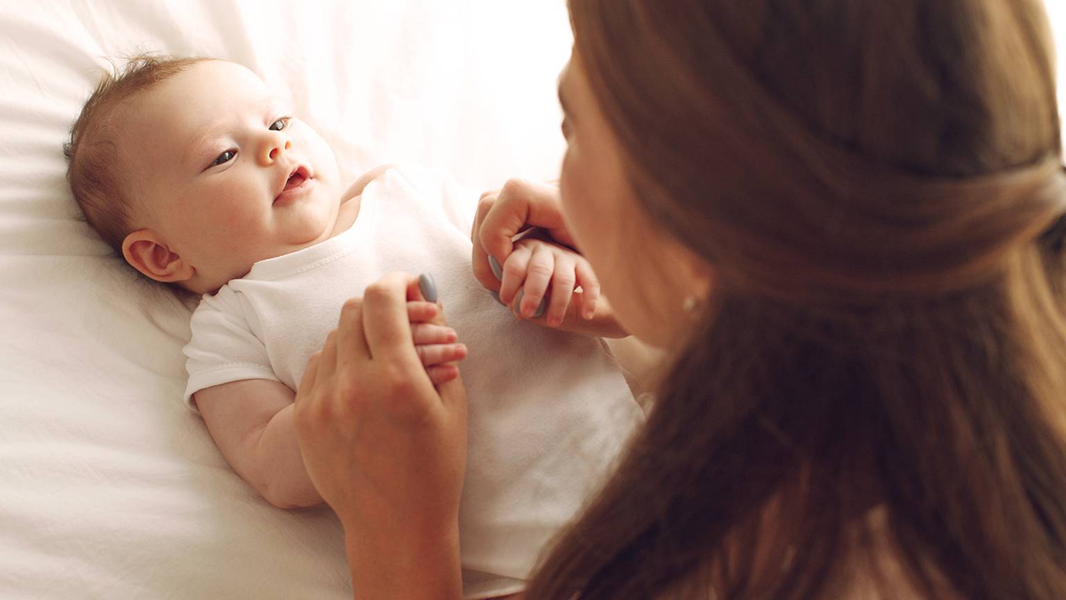 Eine Frau hält sanft die Hände eines Babys, während sie auf einem weißen Bett liegen und sich beide mit sanftem Gesichtsausdruck anschauen.