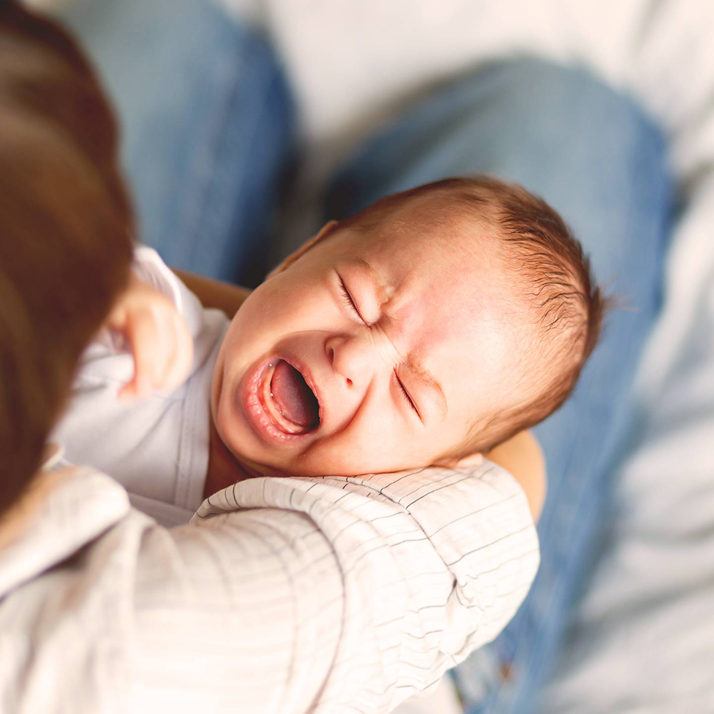 Ein weinendes Baby liegt in den Armen einer Frau und trägt ein weißes Outfit. Der Hintergrund ist leicht unscharf und betont den Gesichtsausdruck des Babys.