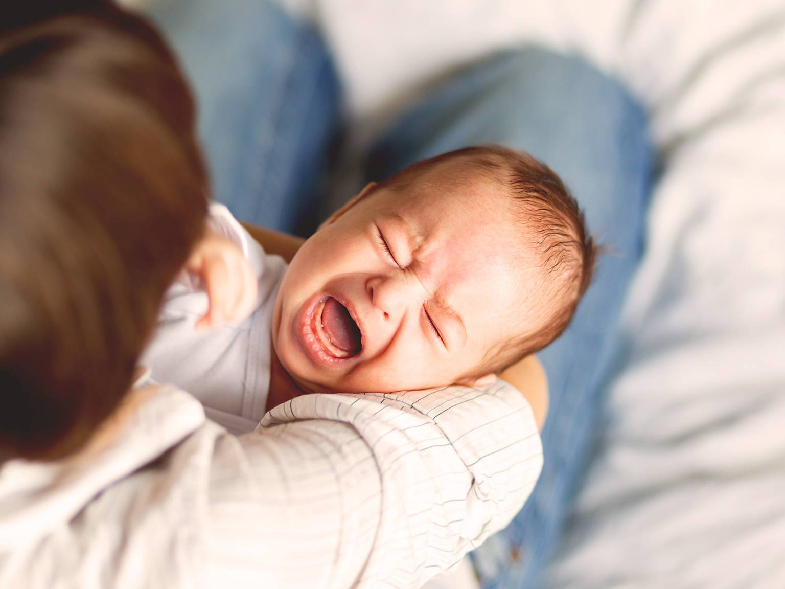 Ein weinendes Baby liegt in den Armen einer Frau und trägt ein weißes Outfit. Der Hintergrund ist leicht unscharf und betont den Gesichtsausdruck des Babys.