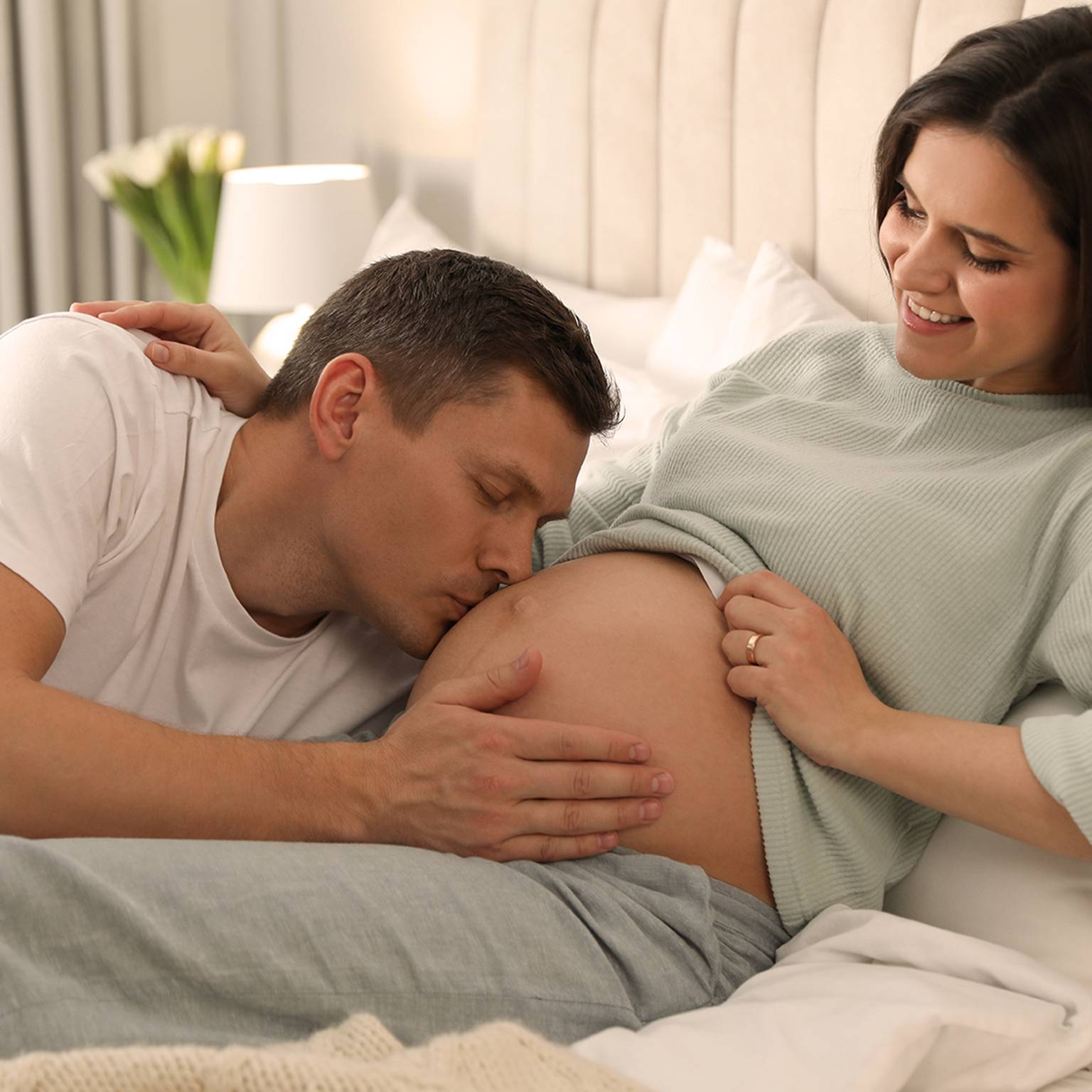 Ein Mann küsst sanft den Bauch einer schwangeren Frau, während sie lächelt und auf einem Bett in einem gemütlichen, sanft beleuchteten Raum sitzt.