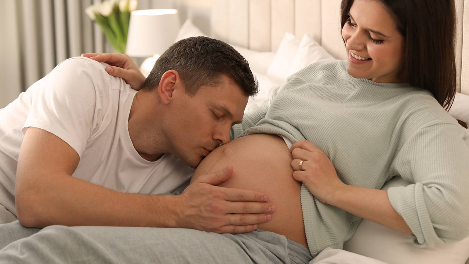 Ein Mann küsst sanft den Bauch einer schwangeren Frau, während sie lächelt und auf einem Bett in einem gemütlichen, sanft beleuchteten Raum sitzt.