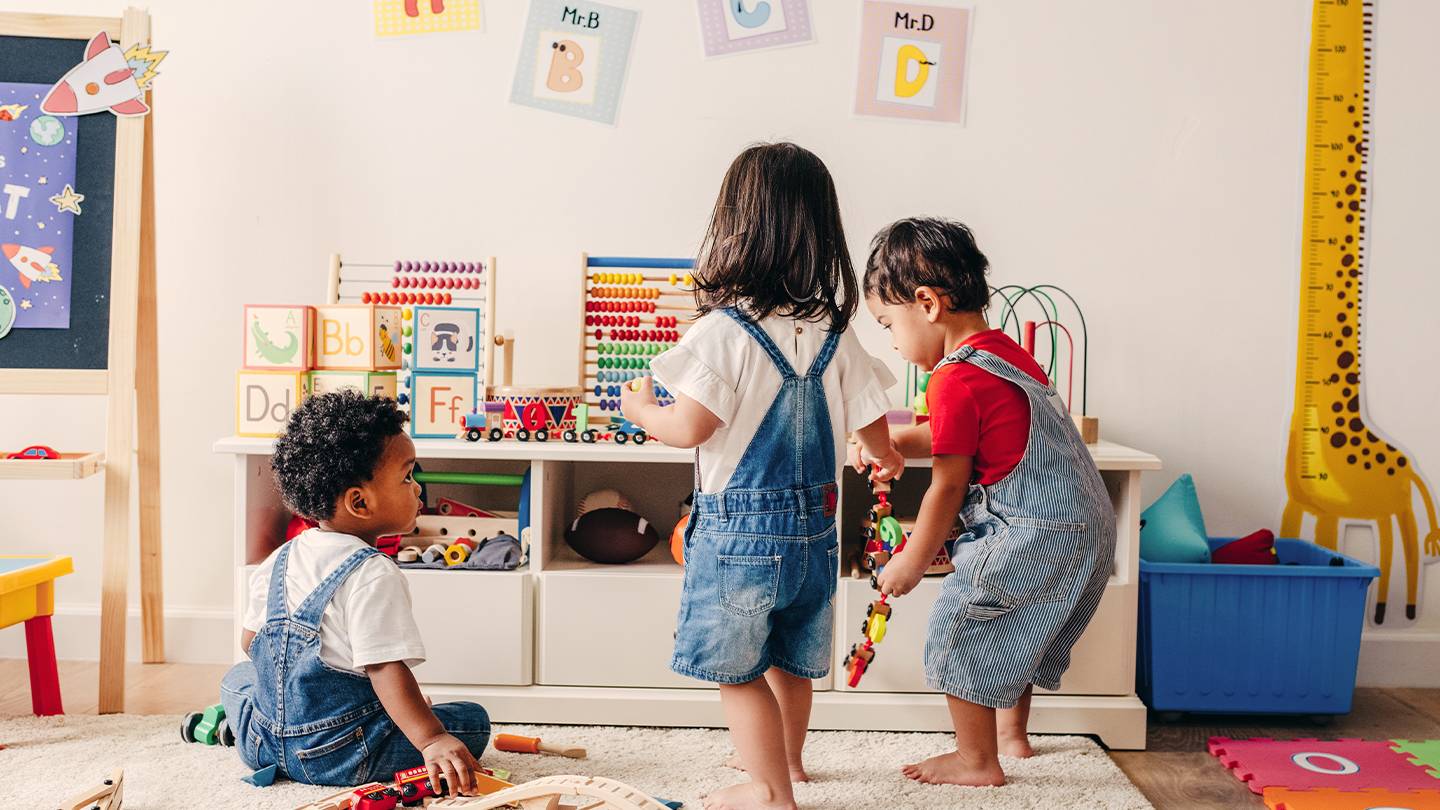 Drei Kinder in Jeans-Overalls mit Spielzeug in einem Spielzimmer. Alphabet-Poster und bunte Dekorationen schmücken die Wände.