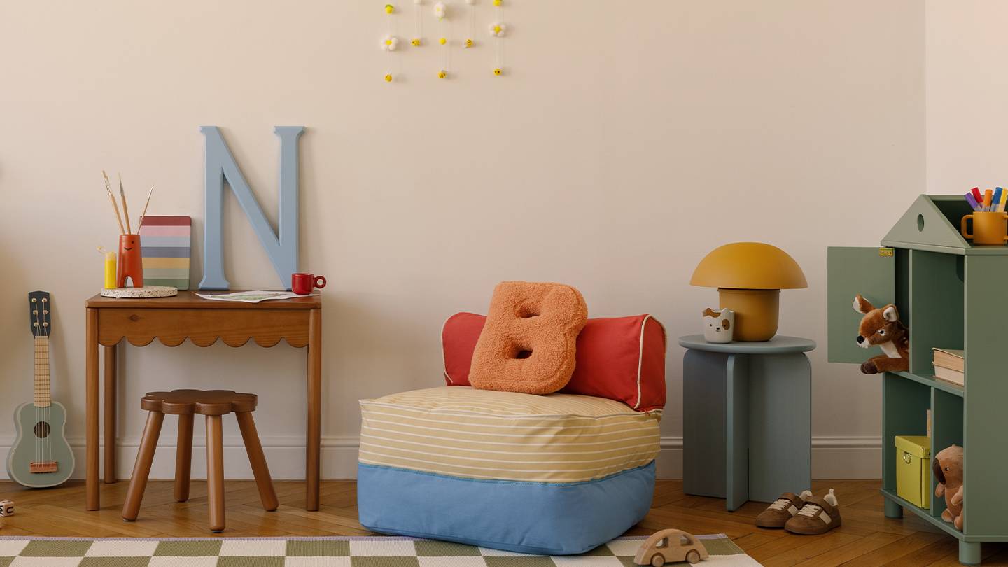 Gemütliches Kinderzimmer mit kleinem Schreibtisch, Sitzsack, Spielsachen und Dekoartikeln. Ein karierter Teppich bedeckt den Holzboden.