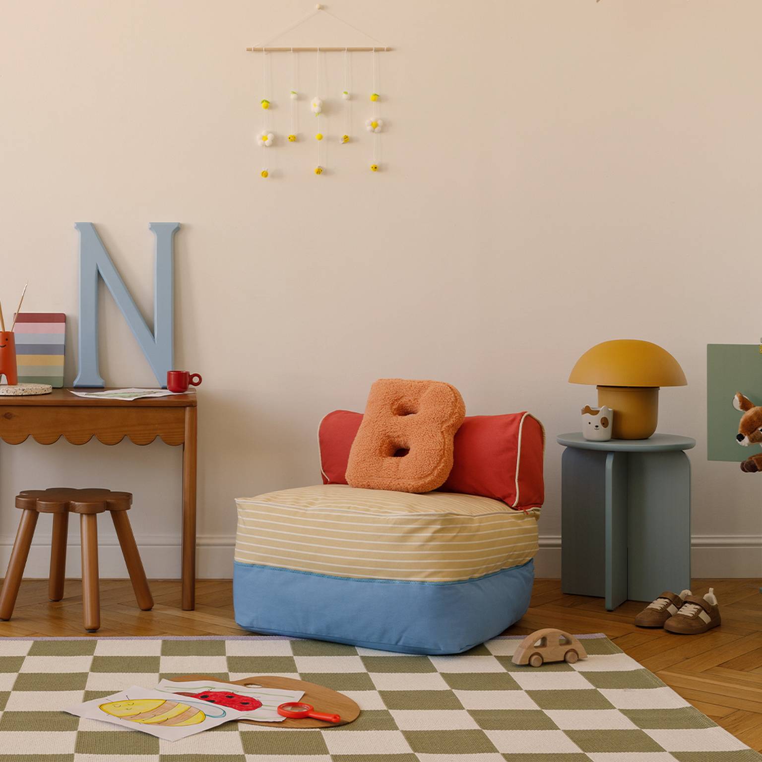 Gemütliches Kinderzimmer mit kleinem Schreibtisch, Sitzsack, Spielsachen und Dekoartikeln. Ein karierter Teppich bedeckt den Holzboden.