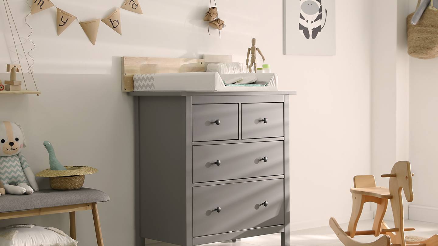 Ein gemütliches Kinderzimmer mit grauem Wickeltisch, Wanddeko, Schaukelpferd und Kuscheltieren. Neutrale Töne schaffen eine beruhigende Atmosphäre.