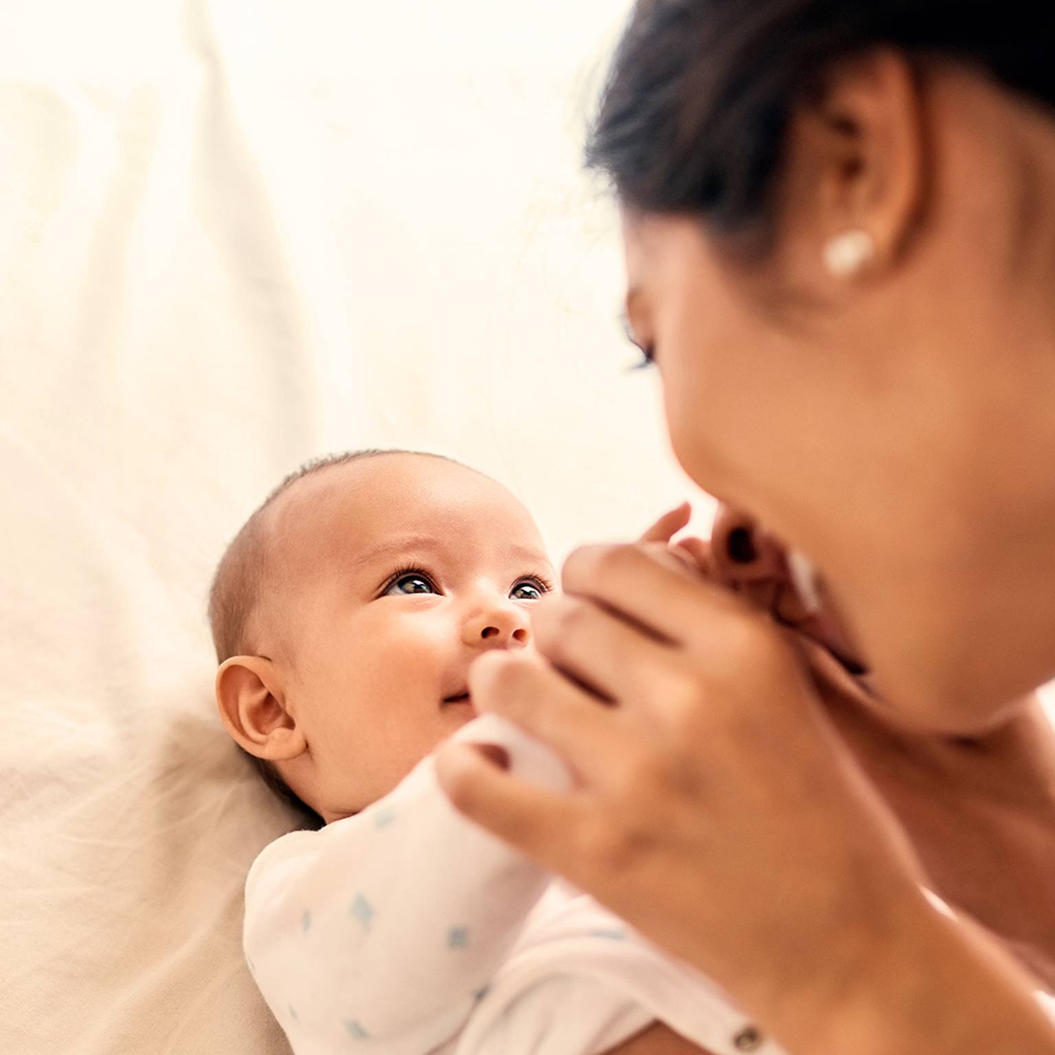 Ein lächelndes Baby, das auf einem Bett liegt, blickt zu einer Frau auf, die sanft die Hände des Babys hält, wodurch eine warme und freudige Interaktion entsteht.
