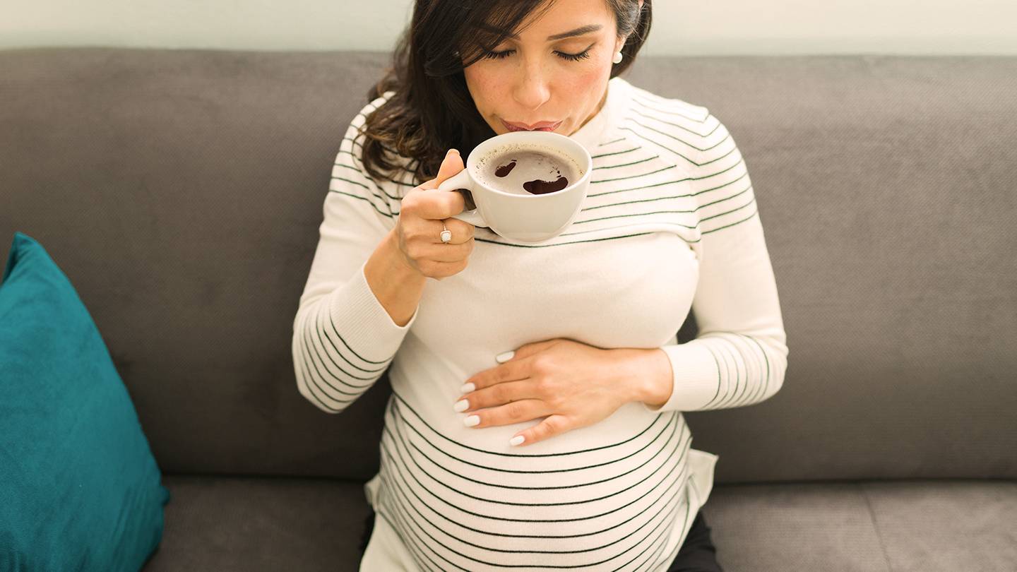 Eine schwangere Frau in einem gestreiften Oberteil sitzt auf einem Sofa, hält ihren Bauch und nippt an einer weißen Tasse, neben ihr liegt ein blaugrünes Kissen.