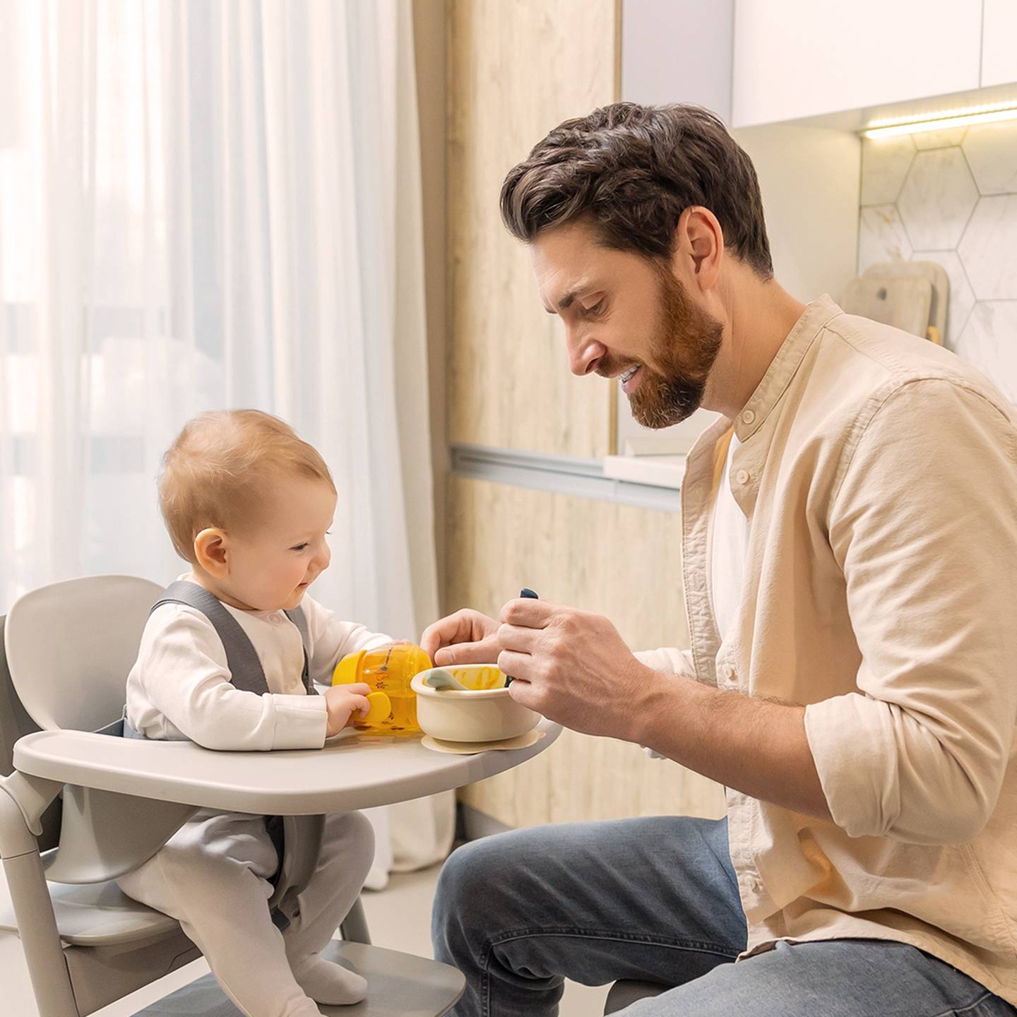 Vater füttert Baby zu Hause im Hochstuhl. Das Baby hält ein Spielzeug, während der Vater ihm einen Löffel Essen aus einer Schüssel anbietet.